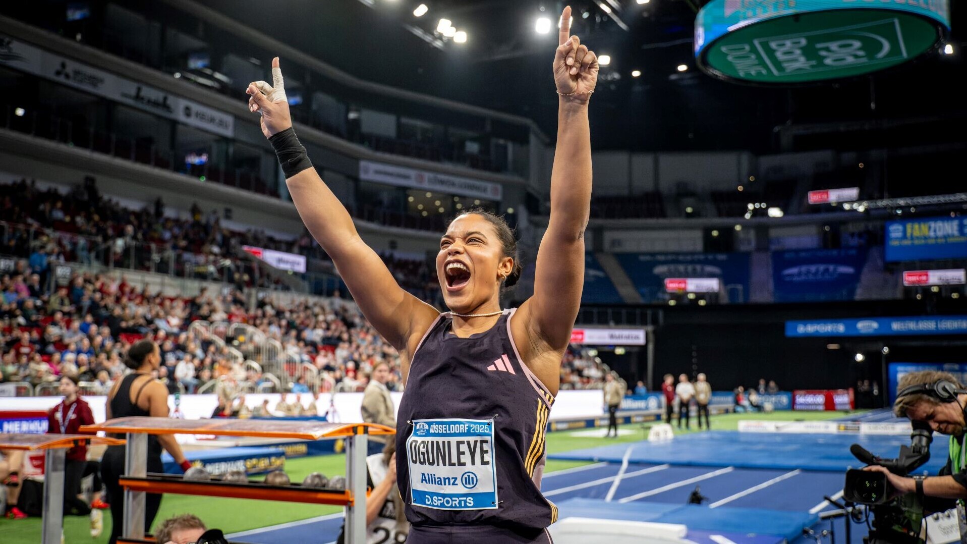Yemisi Ogunleye strahlt vor der Kugelstoß-Anlage in der Halle und reckt die Hände in die Höhe 