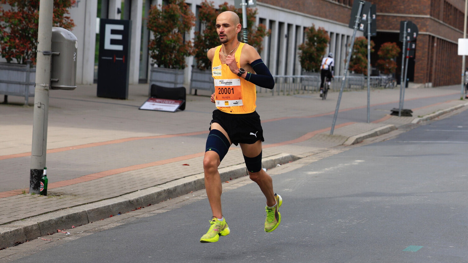 Der deutsche Langstreckler Hendrik Pfeiffer läuft mit der Startnummer 20001 eine leere Stadtstraße entlang. Er trägt ein orangefarbenes Trägershirt, schwarze Shorts, Kompressionsstrümpfe und neongelbe Laufschuhe. Im Hintergrund stehen ein Backsteingebäude, Bäume und ein Radfahrer.