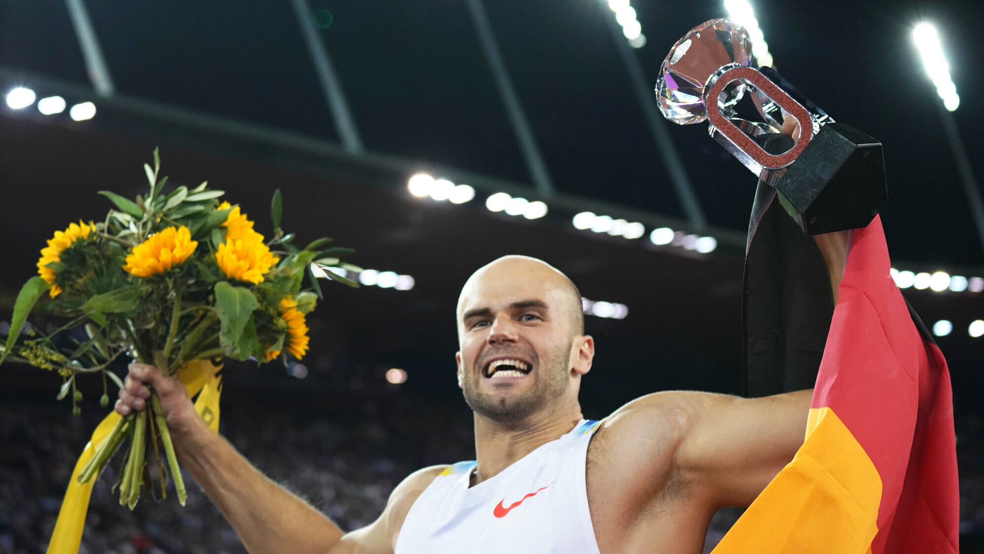 Speerwerfer Julian Weber reckt in Zürich 2025 jubelnd den Diamond-League-Pokal mit dem großen Kristall in die Höhe. In der anderen Hand hält er einen Sonnenblumenstrauß, über seiner Schulter hängt eine Deutschlandfahne. Im Hintergrund ist das nächtliche Stadion mit Flutlicht zu sehen.