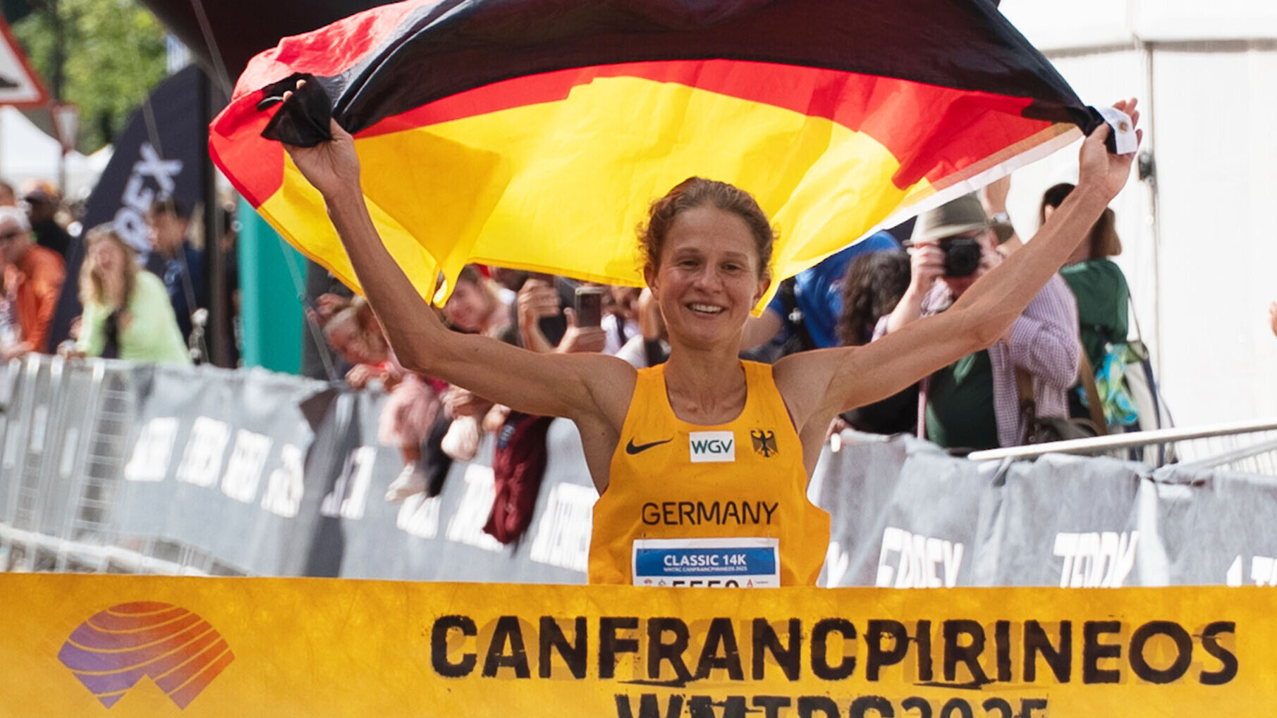 Nina Engelhard hält beim Zieleinlauf bei der Berg- und Traillauf-WM 2025 in Canfranc jubelnd eine Deutschlandfahne über den Kopf. Sie trägt das gelbe Deutschland-Trikot, im Hintergrund jubeln Zuschauende.