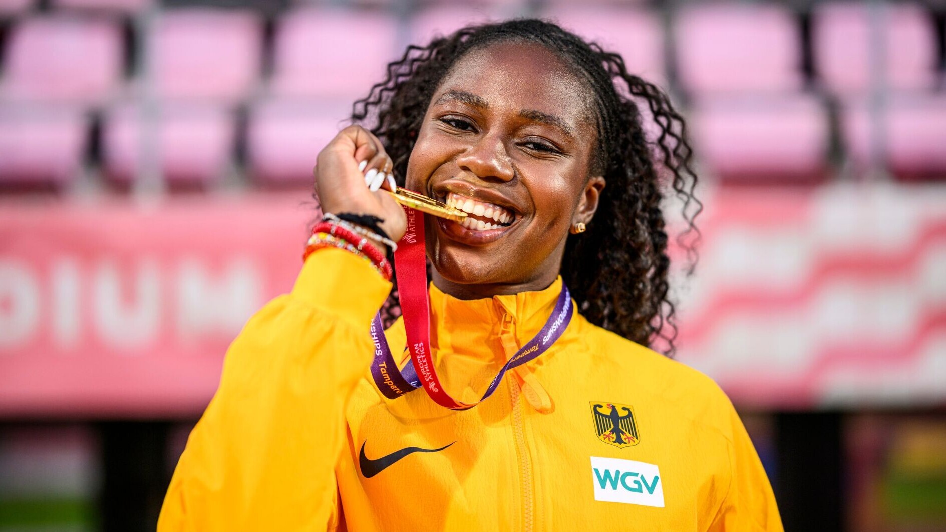 Judith Mokobe beißt lachend auf eine Goldmedaille. Sie trägt eine gelbe Jacke der deutschen Nationalmannschaft mit Bundesadler, dahinter sind leere pinkfarbene Tribünensitze zu sehen. Auf dem Medaillenband steht „Tampere“.