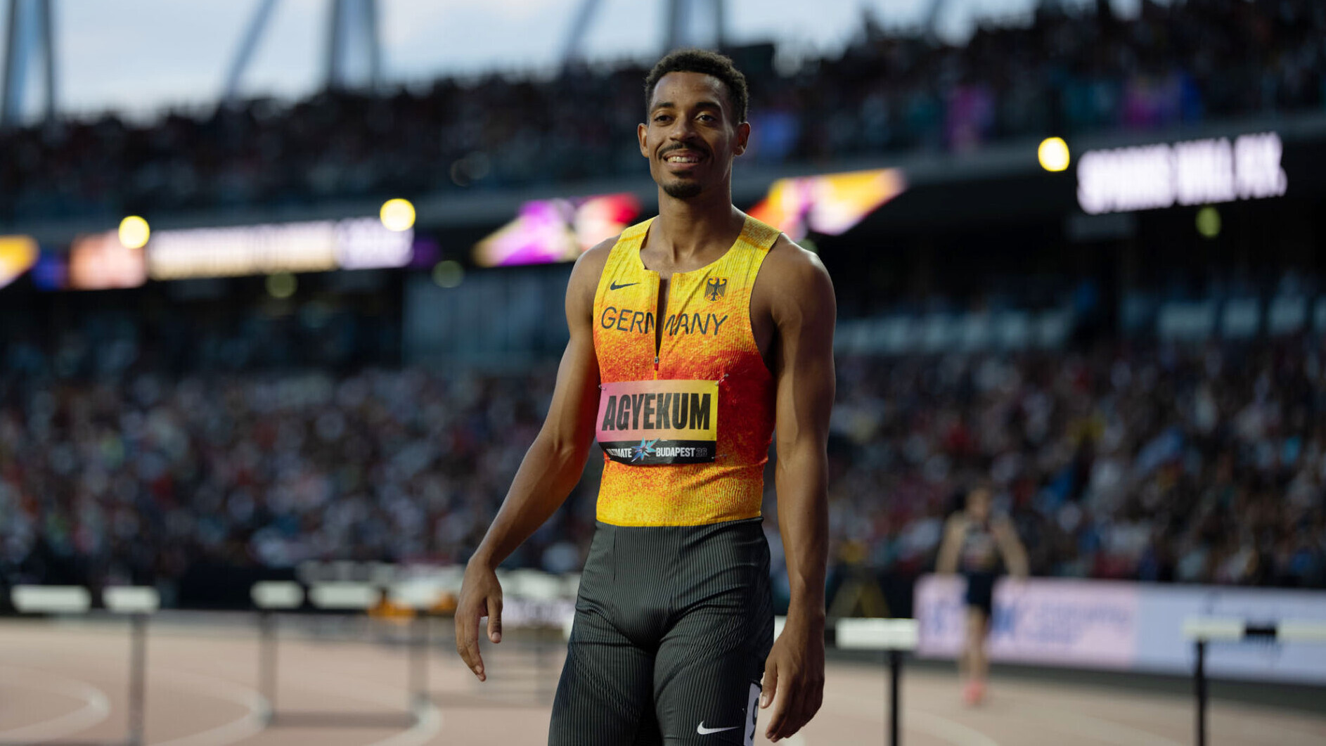 Der deutsche Leichtathlet Emil Agyekum steht im Nationaltrikot vor einem Rennen in einem Stadion und lächelt in die Kamera.