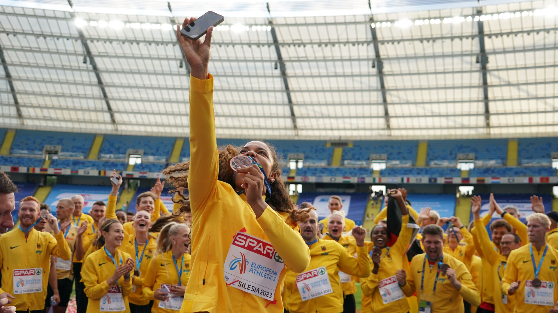 Kugelstoßerin Yemisi Ogunleye macht bei der Team-EM 2023 ein Selfie mit sich und dem deutschen Team im Hintergrund, alle tragen gelbe Trainingsanzüge