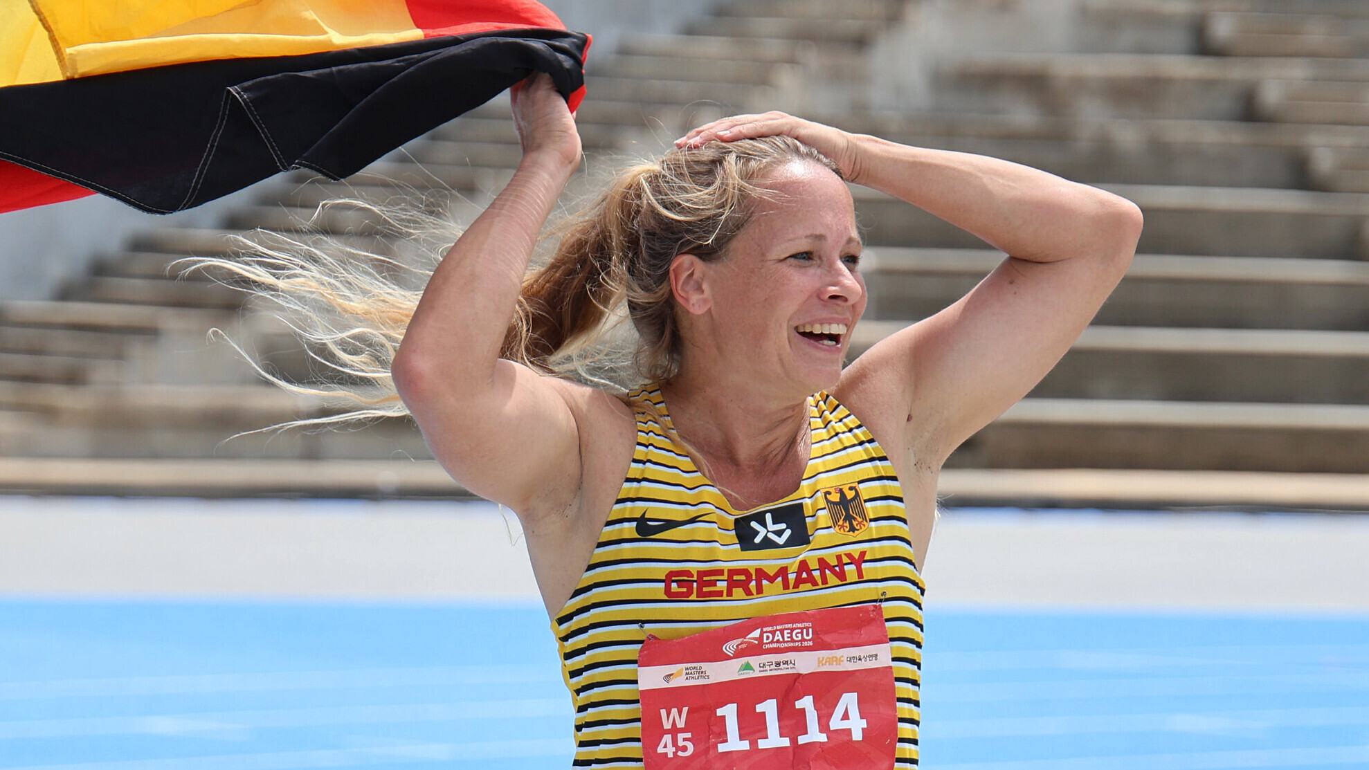 Die deutsche Masters-Athletin Mareike Metz jubelt mit den Armen über dem Kopf und trägt eine Deutschlandfahne in der rechten Hand.