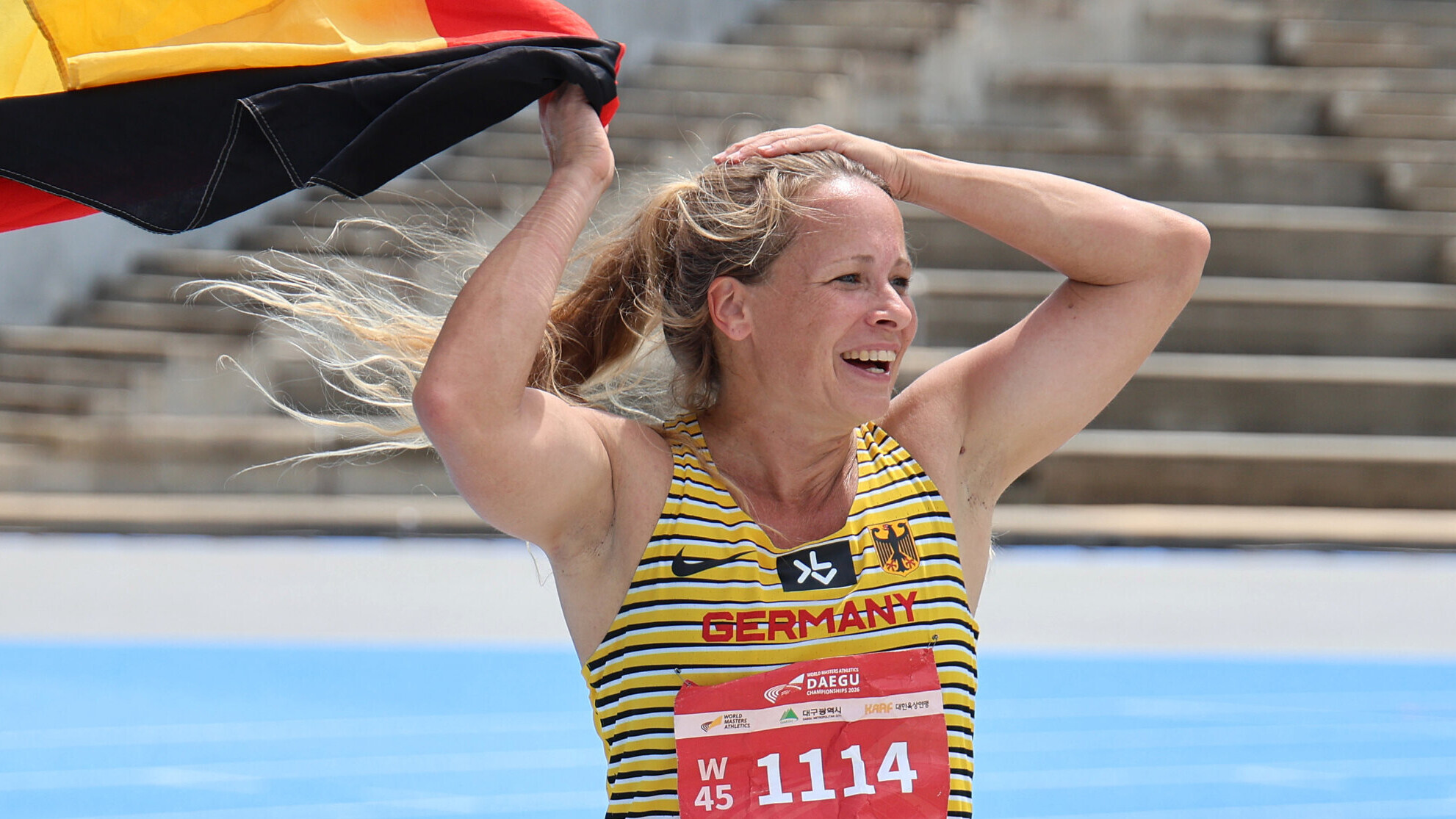 Die deutsche Masters-Athletin Mareike Metz jubelt mit den Armen über dem Kopf und trägt eine Deutschlandfahne in der rechten Hand.