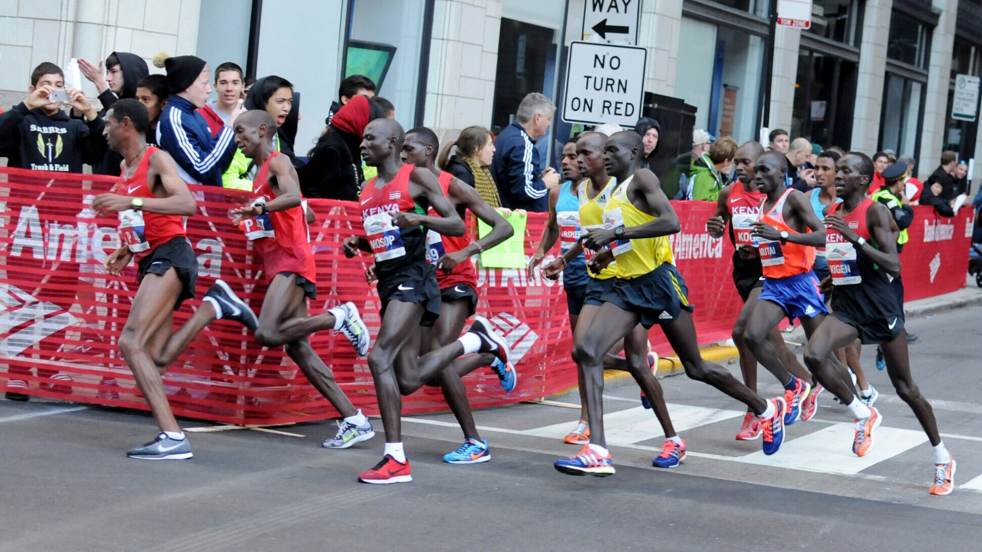 Eine Spitzengruppe von rund zehn Läufern beim Chicago-Marathon, viele davon in kenianischen Trikots, läuft eine Großstadtstraße entlang. Hinter einer roten Absperrung mit Bank-of-America-Logos feuern Zuschauende sie an.