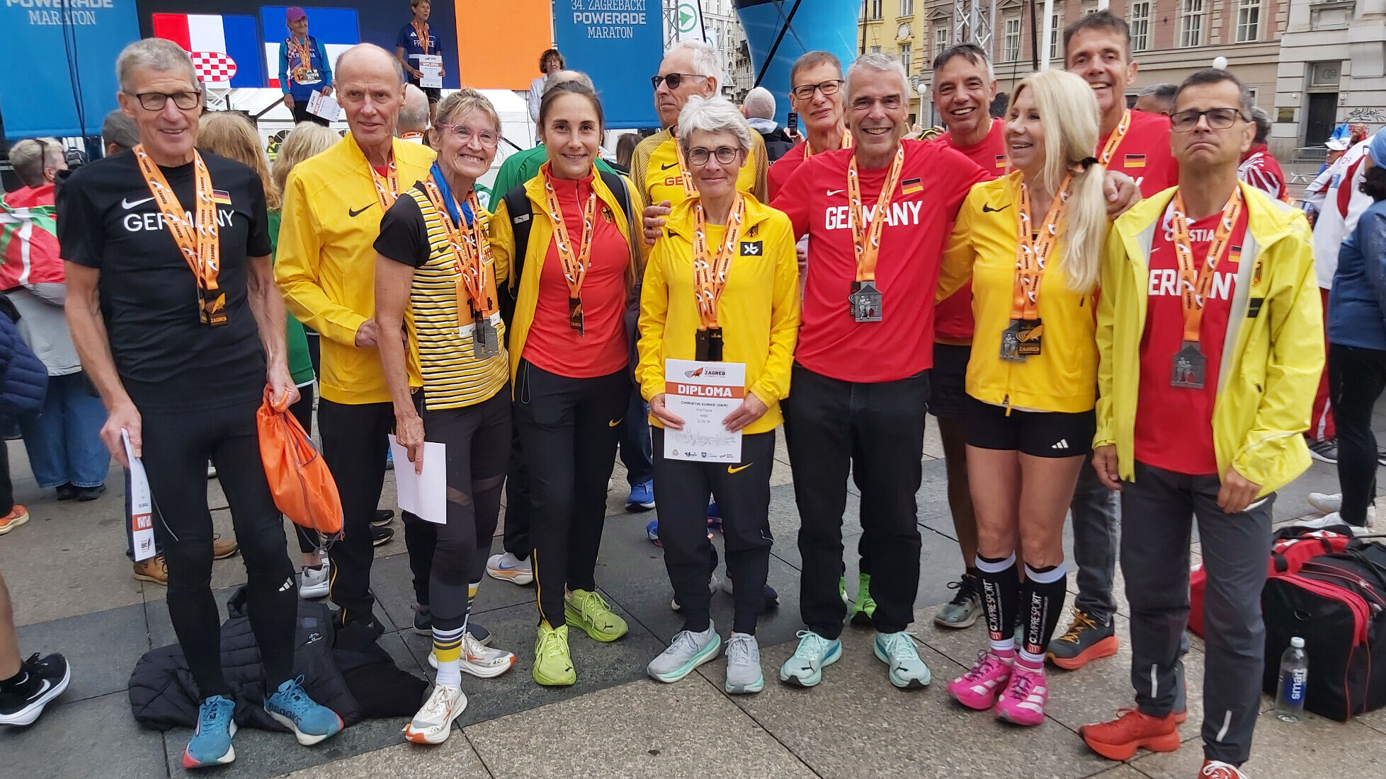 Rund 15 Athletinnen und Athleten des deutschen Masters-Teams stehen nach den Siegerehrungen bei der Marathon-WM in Zagreb gemeinsam für ein Gruppenfoto zusammen. Sie tragen rote, gelbe und schwarze Teamkleidung und Medaillen um den Hals, einige halten Urkunden in den Händen.