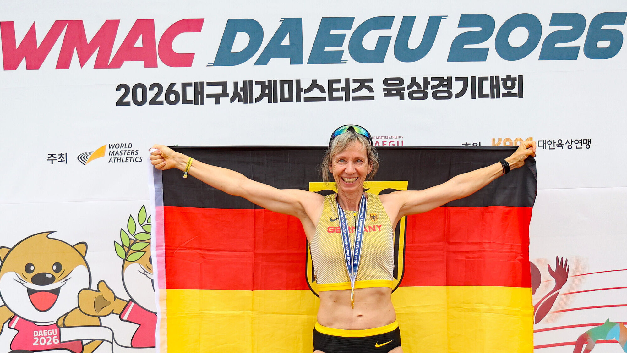 Die deutsche Masters-Athletin Iris Opitz jubelt mit einer Deutschland-Fahne hinter dem Rücken und einer Goldmedaille um den Hals. Im Hintergrund ist der Schritzug WMAC Daegu 2026 zu lesen. 