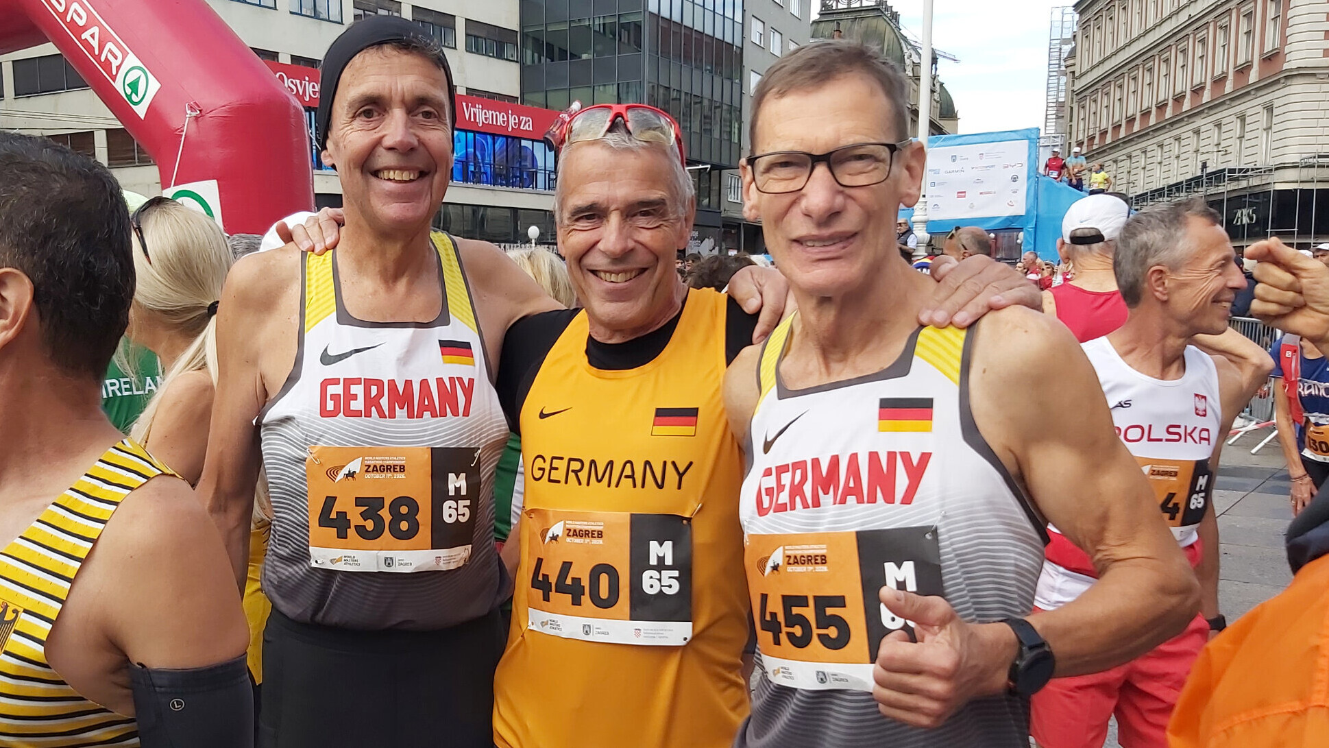 Hermann-Josef Belke, Paul Bückert und Martin Rapp (von links) stehen als deutsches M65-Team Arm in Arm vor dem Start. Sie tragen Deutschland-Trikots mit den Startnummern 438, 440 und 455, im Hintergrund stehen weitere Läuferinnen und Läufer.