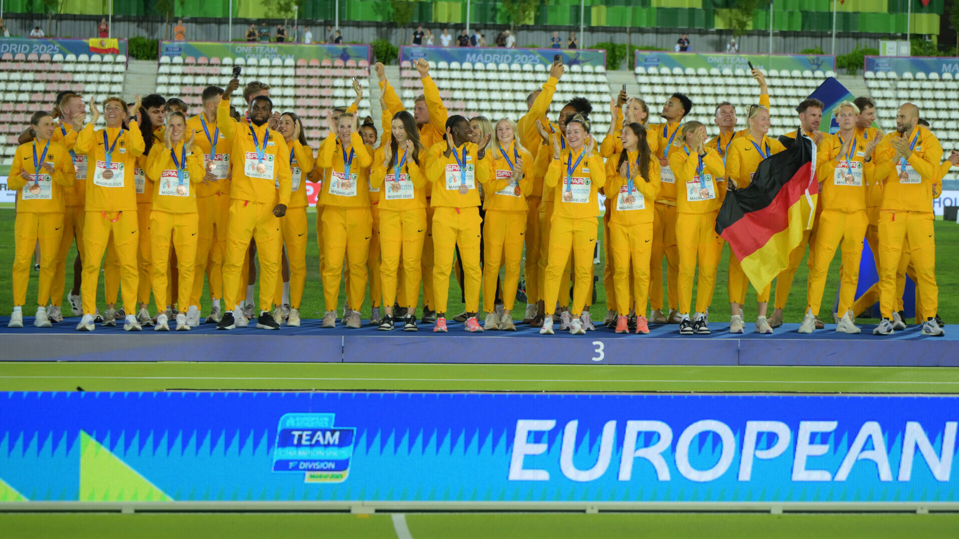 Das deutsche Team jubelt mit Deutschlandfahne und im gelben Podiumsoutfit auf dem Podest der Team-EM in Madrid
