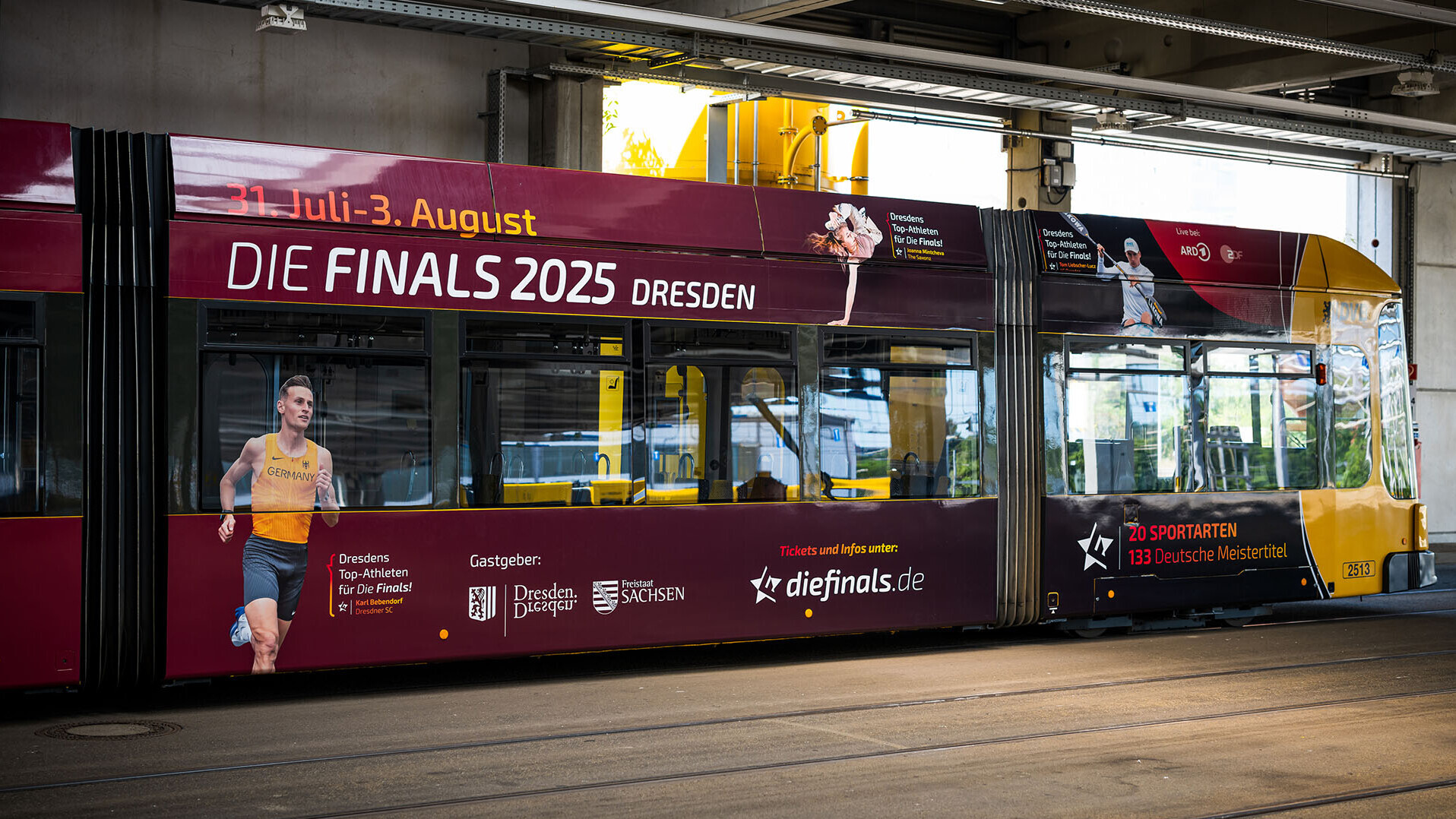 Eine in den Farben schwarz, rot und gold gestaltete Straßenbahn, bedruckt mit Fotos von Athletinnen und Athleten, darunter Hindernisläufer Karl Bebendorf