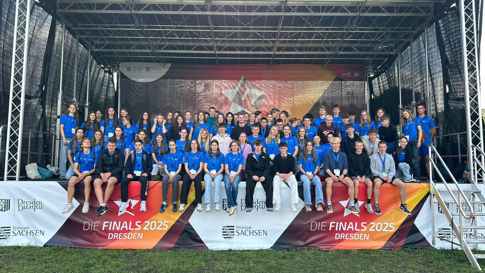 Die Teilnehmenden des DLV-Jugendlagers sitzen, überwiegend in blauen T-Shirts, auf einer Bühne, vor der ein Banner der "Finals 2025 Dresden" hängt