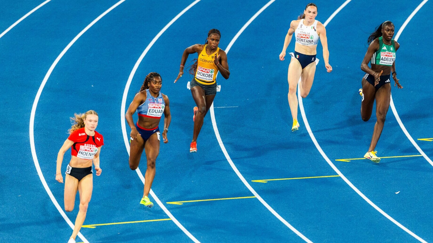 Judith Mokobe läuft im gelben Deutschland-Trikot auf einer blauen Bahn durch die Kurve. Neben ihr laufen Sprinterinnen aus der Schweiz, Großbritannien, Italien und Irland. Die Aufnahme ist von schräg oben gemacht.