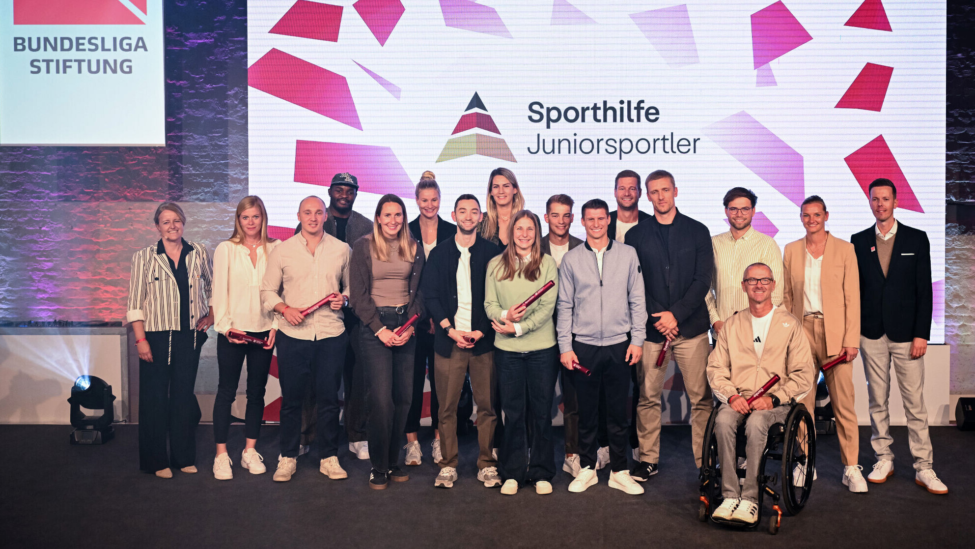 Rund 18 Personen stehen auf einer Bühne bei der Verabschiedung von Athletinnen und Athleten nach ihrer Karriere, eine Person sitzt im Rollstuhl. Viele halten rote Staffelstäbe mit der Aufschrift „Danke“. Auf der Leinwand dahinter steht „Sporthilfe Juniorsportler“, links hängt ein Schild der Bundesliga-Stiftung.