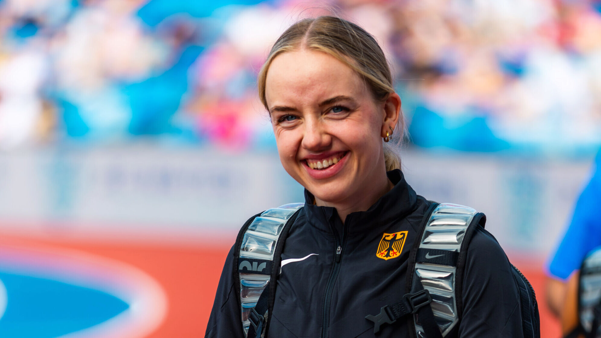 Die deutsche Weitspringerin Imke Daalmann lächelt in die Kamera. Sie trägt eine schwarze Trainingsjacke der Nationalmannschaft mit Bundesadler und einen Rucksack, im Hintergrund ist unscharf ein voll besetztes Stadion zu sehen.
