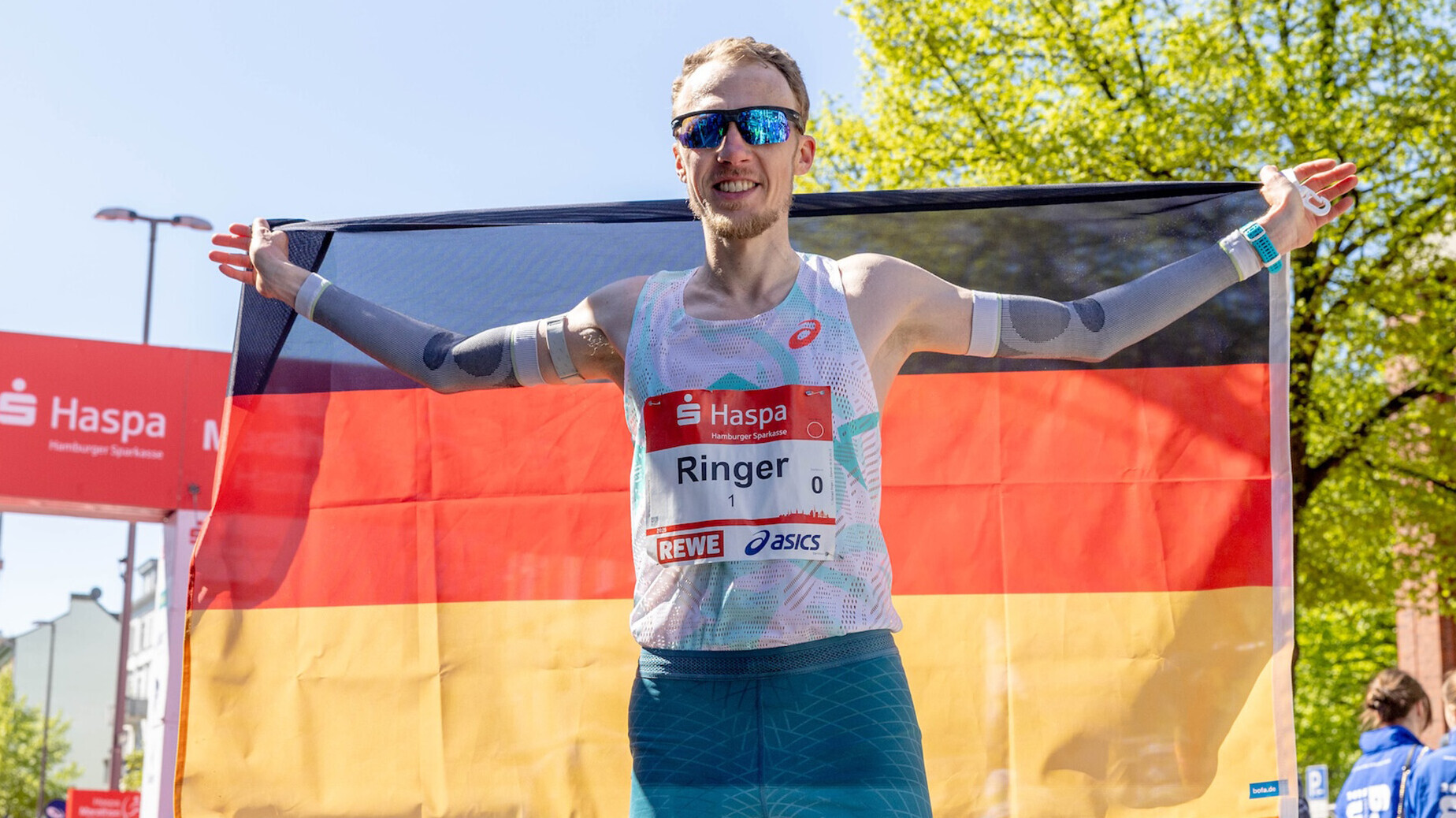 Der deutsche Marathonläuer Richard Ringer lächelt in die Kamera und hält dabei eine Deutschlandfahne hinter dem Rücken.