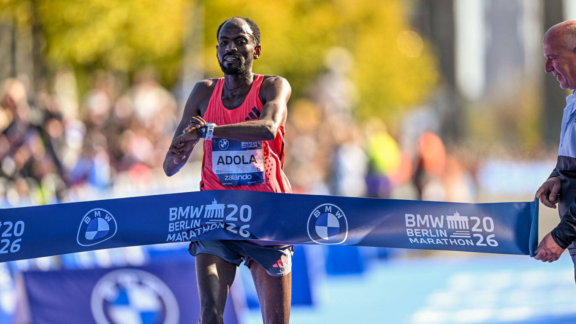 Der äthiopische Langstreckler Guye Adola durchläuft ein Zielband mit der Aufschrit "Berlin-Marathon 2026" und drückt dabei auf seine Uhr.
