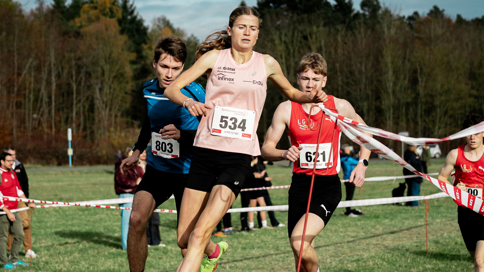Julia Ehrle läuft mit der Startnummer 534 beim Pforzheim-Cross im rosafarbenen Vereinstrikot über eine Wiese bei einem Crosslauf. Neben ihr laufen zwei junge Männer mit den Startnummern 803 und 621, einer davon schiebt rot-weißes Absperrband zur Seite. Im Hintergrund stehen herbstliche Bäume und Zuschauende an der Strecke.
