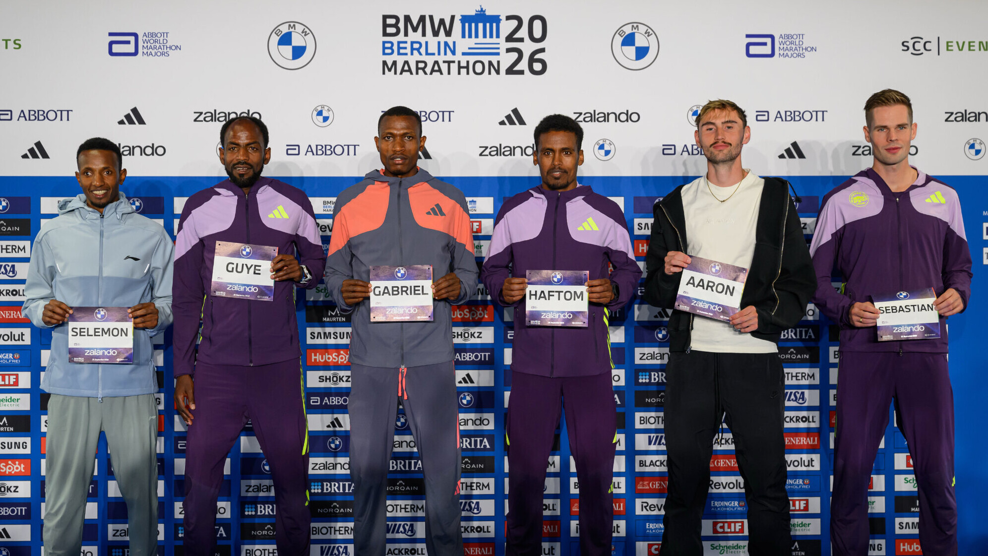 Sechs Läufer der Männer-Elite stehen bei der Pressekonferenz zum BMW Berlin-Marathon 2026 vor einer Sponsorenwand und halten Startnummern mit ihrem Vornahmen vor dem Körper: von links Selemon Barega, Guye Adola, Gabriel Geay, Haftom Welday, Aaron Bienenfeld und Sebastian Hendel.