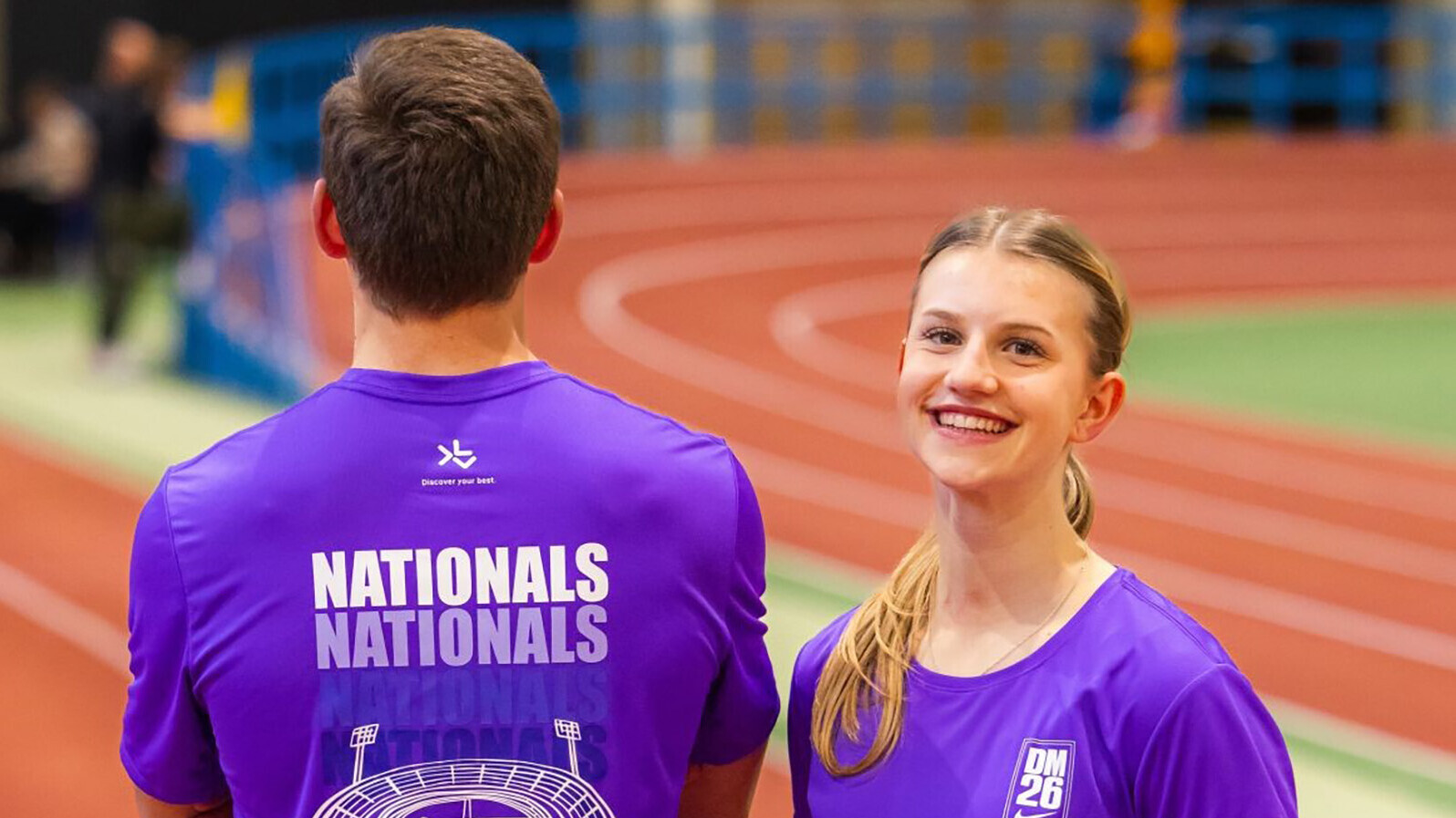 Ein Mann steht mit dem Rücken zur Kamera, eine Frau schaut in die Kamera. Beide tragen ein lilafarbenes Shirt, auf dem vorne DM 2026 steht und hinten "Nationals". Darunter ist ein Stadion abgebildet. 