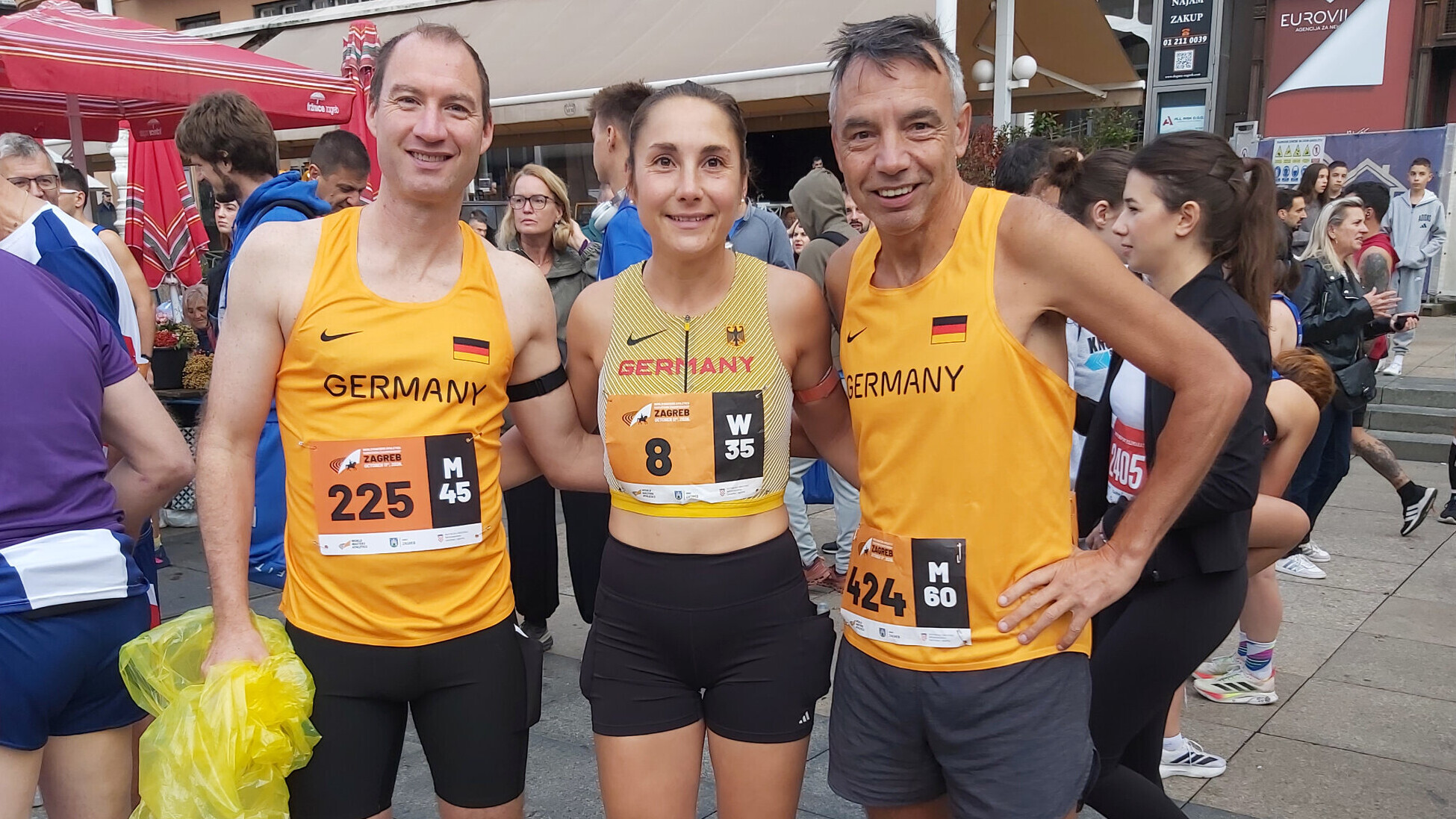 Dirk Joos, Sylvie Müller und Robert Rohregger (von links) stehen in deutschen Trikots lächelnd nebeneinander vor dem Start, im Hintergrund warten weitere Läuferinnen und Läufer.