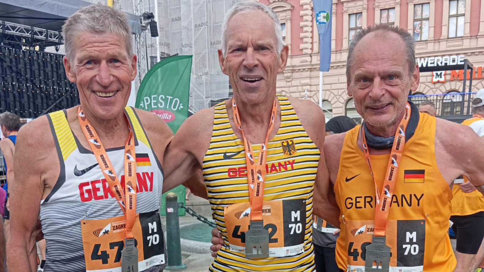 Thomas Jäger (Mitte), Doppelweltmeister der M70, steht lachend zwischen seinen Teamkollegen Peter Massny (links) und Albert-Eugen Vetter (rechts). Alle drei tragen Deutschland-Trikots, Startnummern und Medaillen um den Hals.