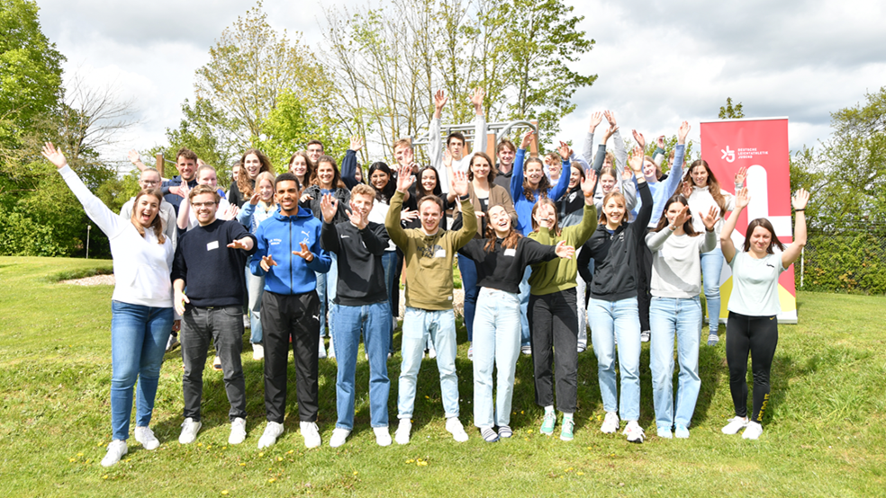 Ein Gruppenbild mit etwa 25 jungen Menschen, die auf einer Wiese die Arme in die Höhe strecken.