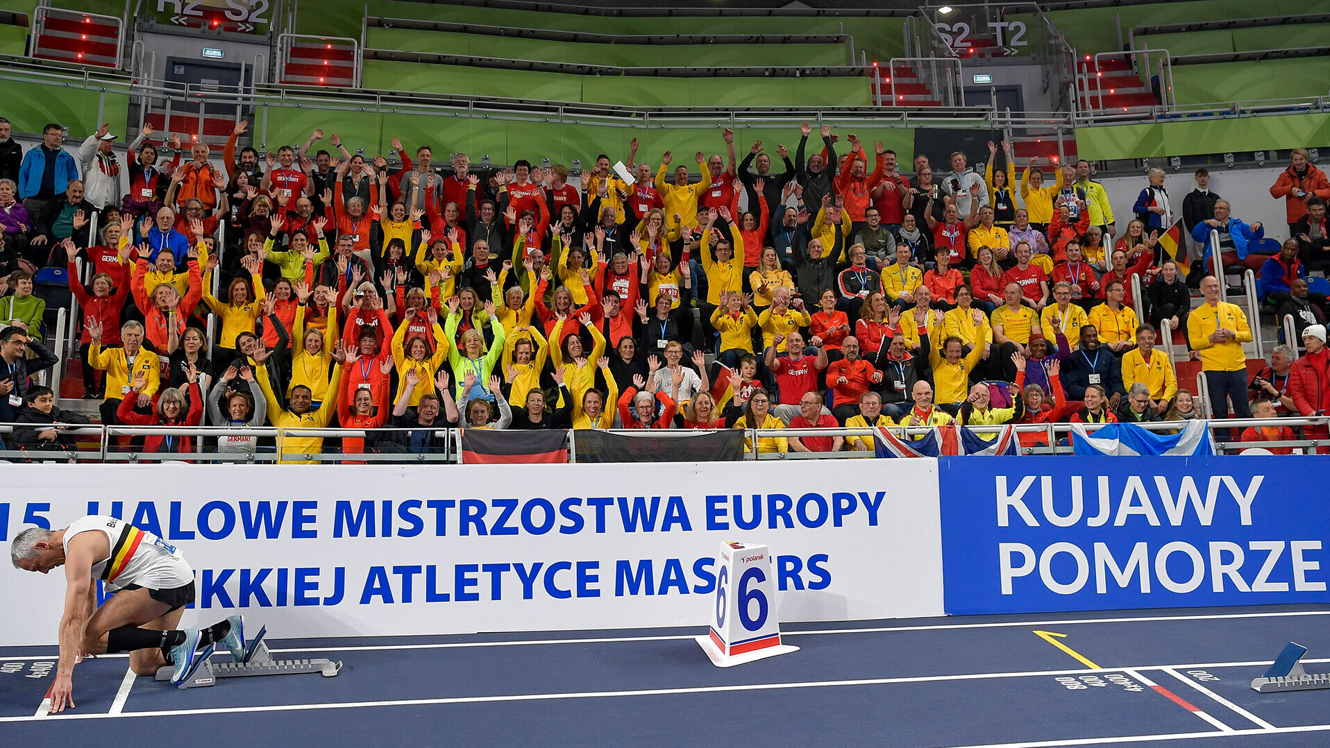 Volle Tribünen mit Personen in roter und gelber Mannschaftskleidung, vorne ein Athlet im Startblock auf der Bahn