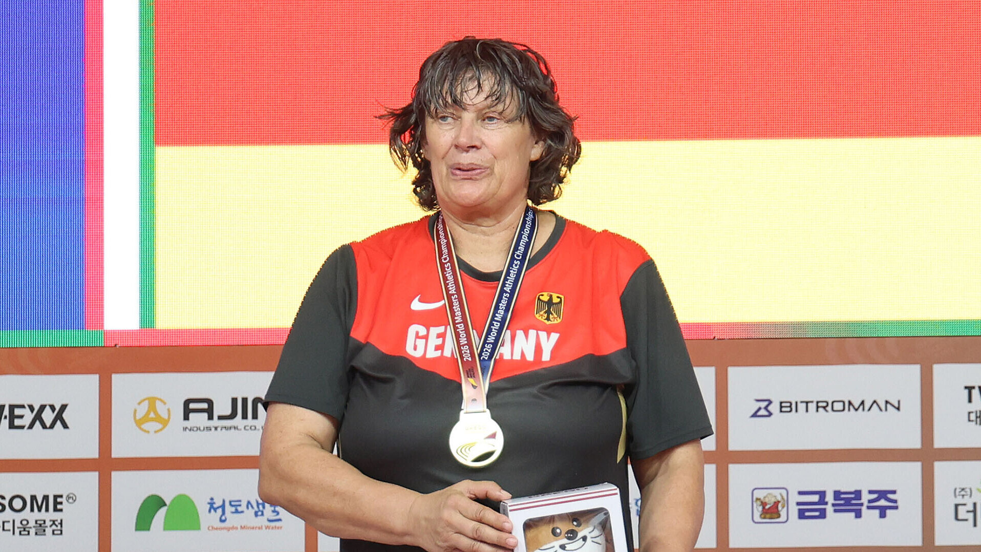 Margret Klein-Raber mit Goldmedaille um den Hals und einem Rahmen in der Hand, im Hintergrund die deutschen Farben