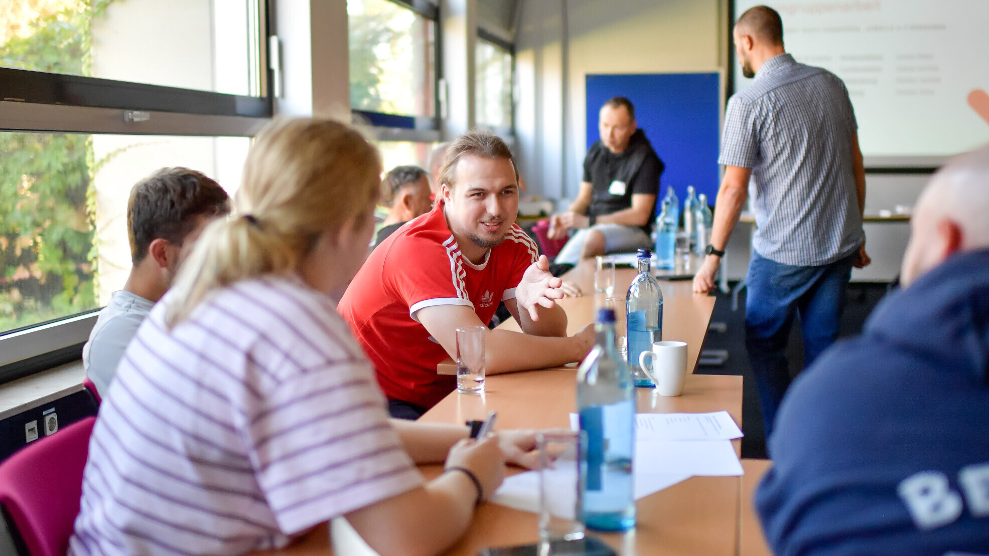 Gruppenarbeit an einer langen Tischreihe: Ein junger Mann im roten Trikot spricht gestikulierend mit Teilnehmenden, im Hintergrund steht ein Referent vor einer Präsentation.