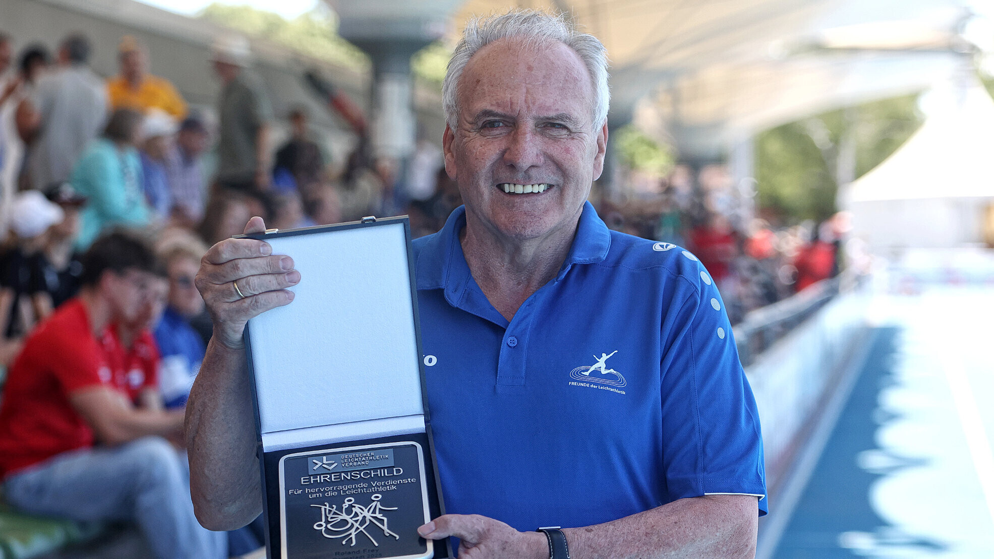 Der Leichtathletik-Funktinör Roland Frey hält die Auszeichnung DLV-Ehrenschild vor dem Körper und lächelt in die Kamera.