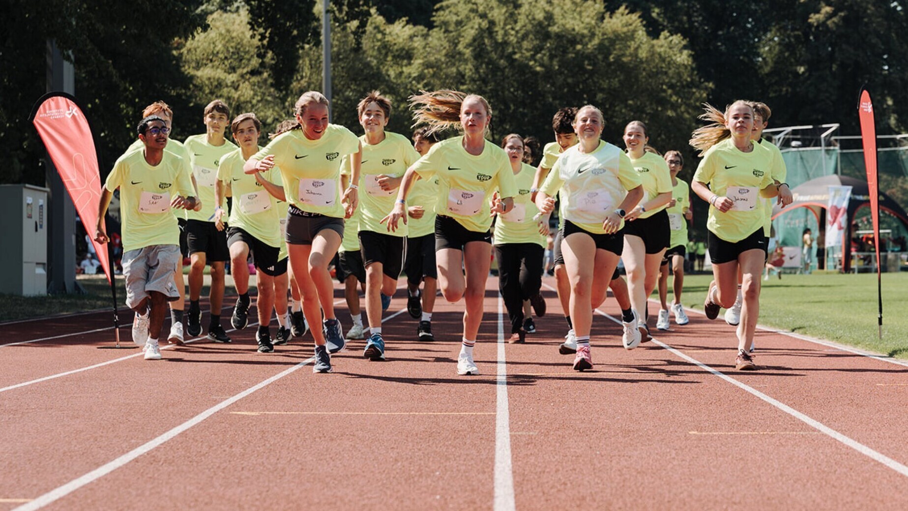 Eine Gruppe Kinder in gelben T-Shirts laufen auf einer roten Laufbahn auf die Kamera zu.