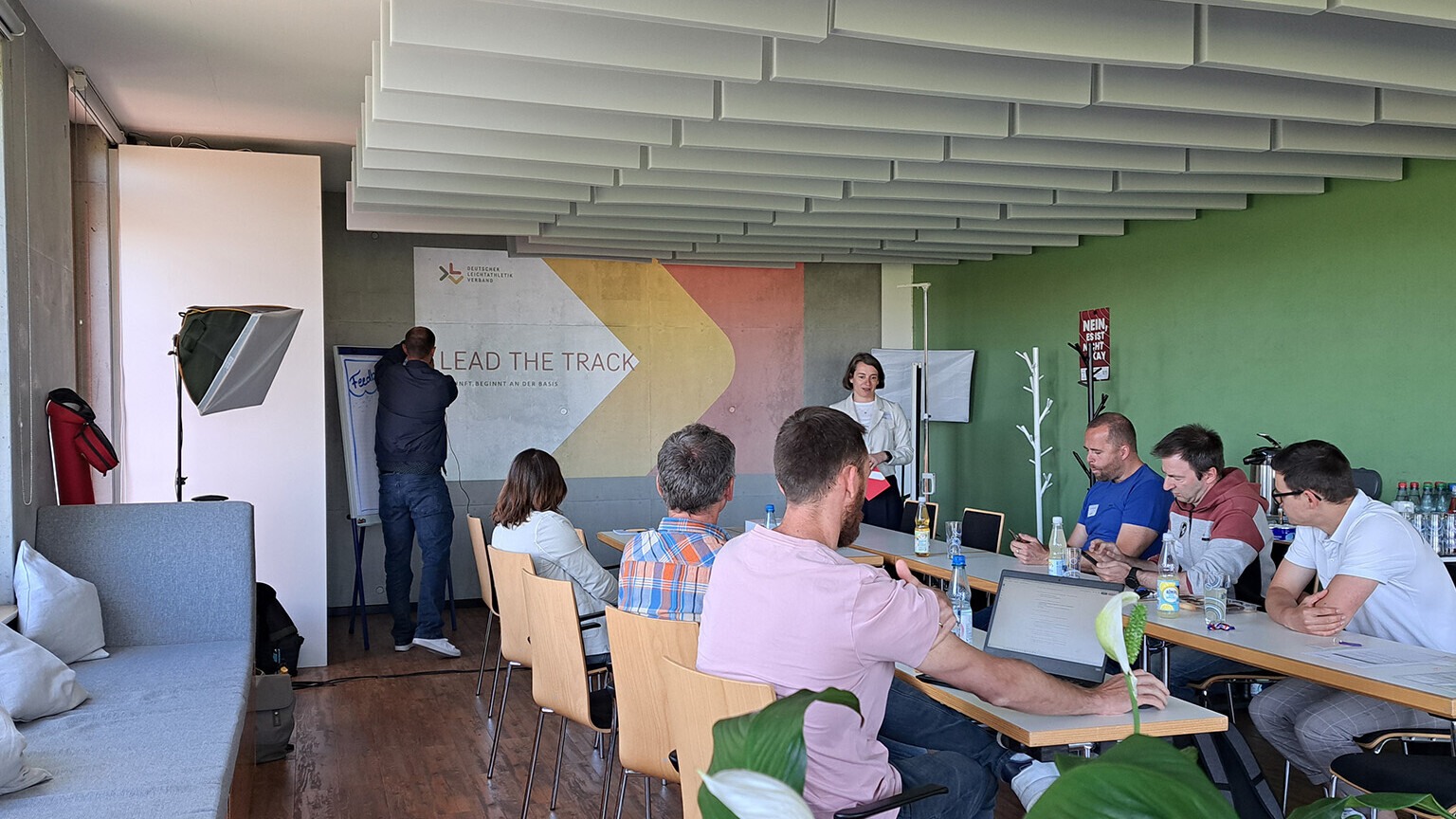 Eine Gruppe Personen an einem Tisch, während Dr. Kristin Behrens vorne steht und eine Präsentation mit dem Titel "Lead the Track" hält.  