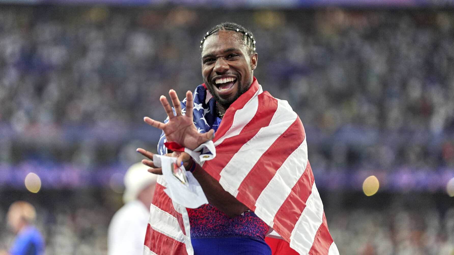 Der US-Sprinter Noah Lyles mit der US-Fahne um den Schultern. Er deutet mit seiner rechten Hand eine Tatze an.