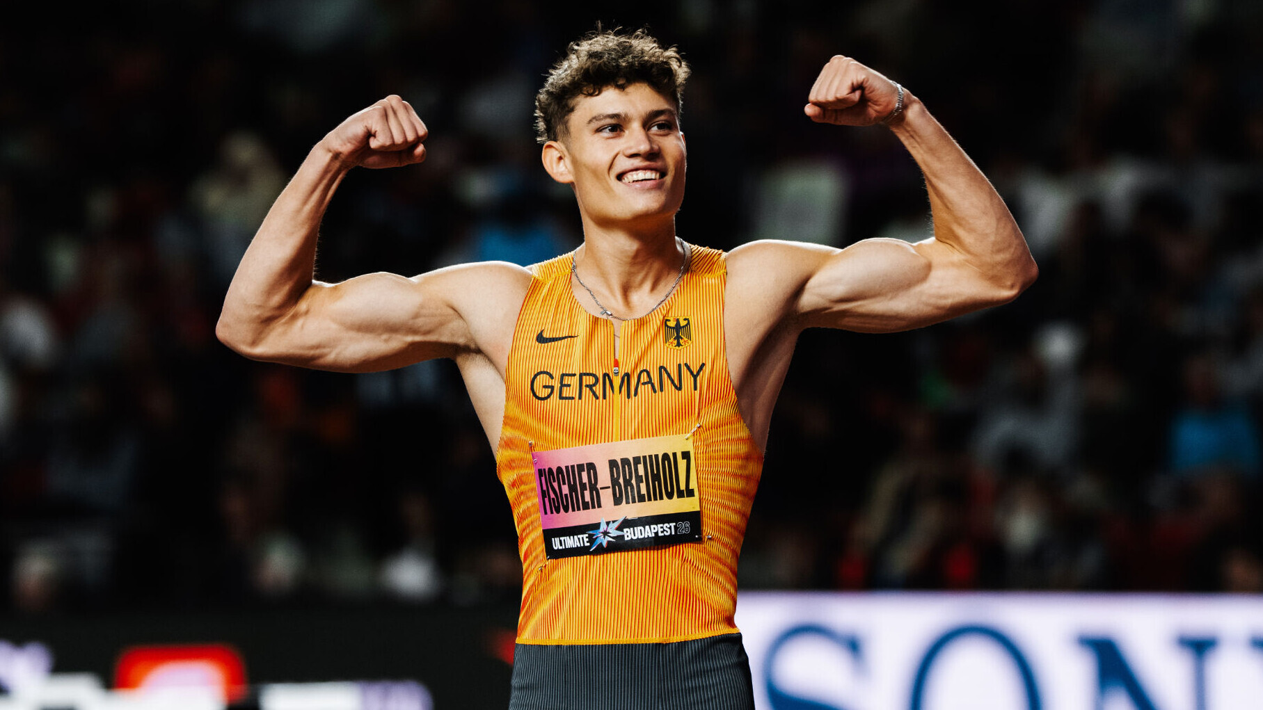 Der deutsche Leichtathlet Owe Fischer-Breiholz hebt im Nationaltrikot die Arme zur Seite und beugt dabei die Arme, sodass der Bizps angespannt ist. Er lächelt dabei in die Kamera.