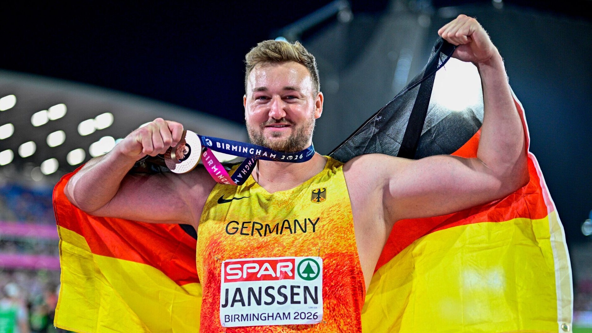 Der Diskuswerfer Henrik Janssen trägt eine Bronzemedaille um den Hals und die Deutschlandfahne um die Schultern, dabei hebt er seine Arme zur Muskelpose.