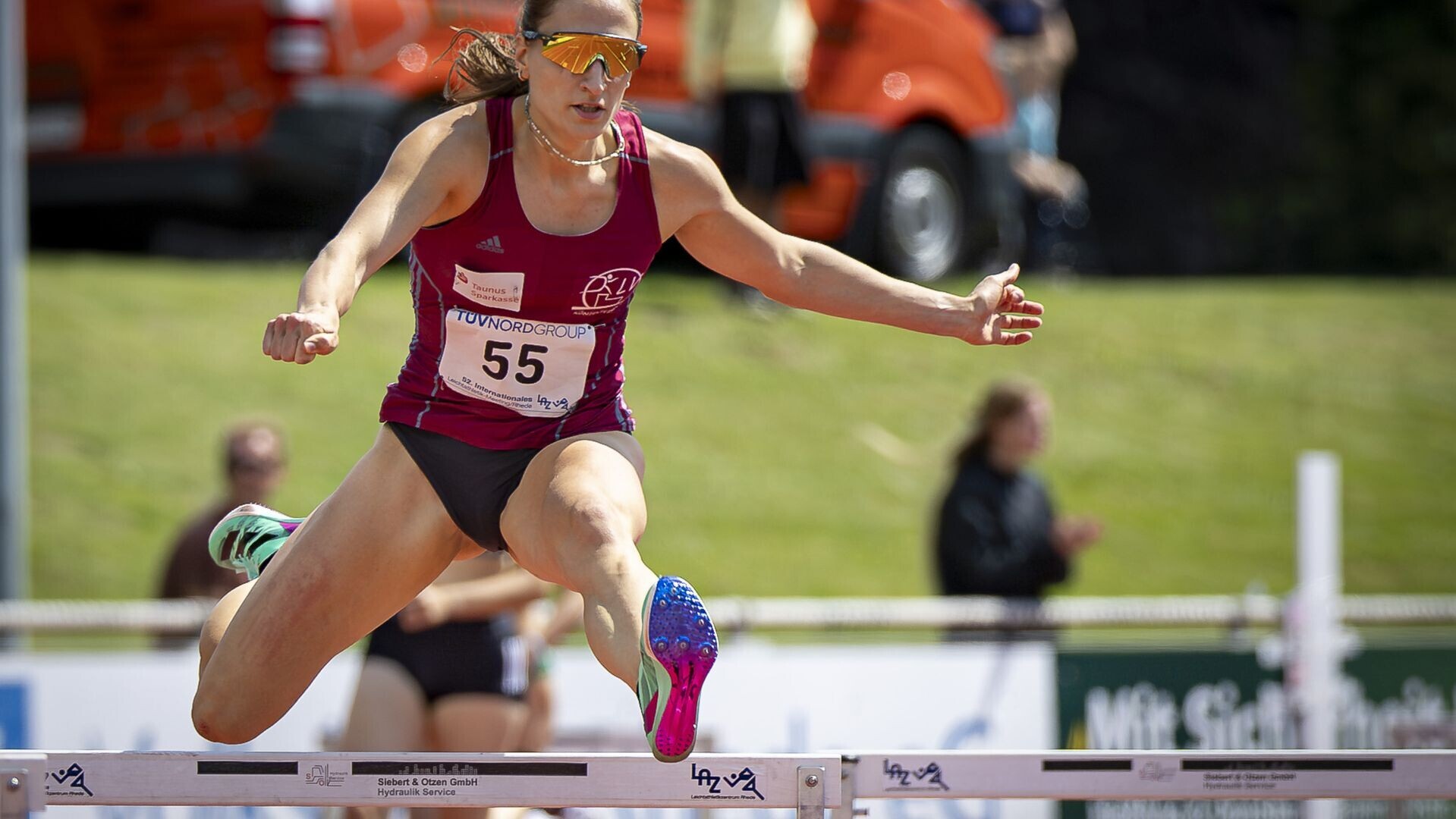 Elena Kelety im roten Vereinstrikot mit Sonnenbrille beim Überqueren einer Hürde. 