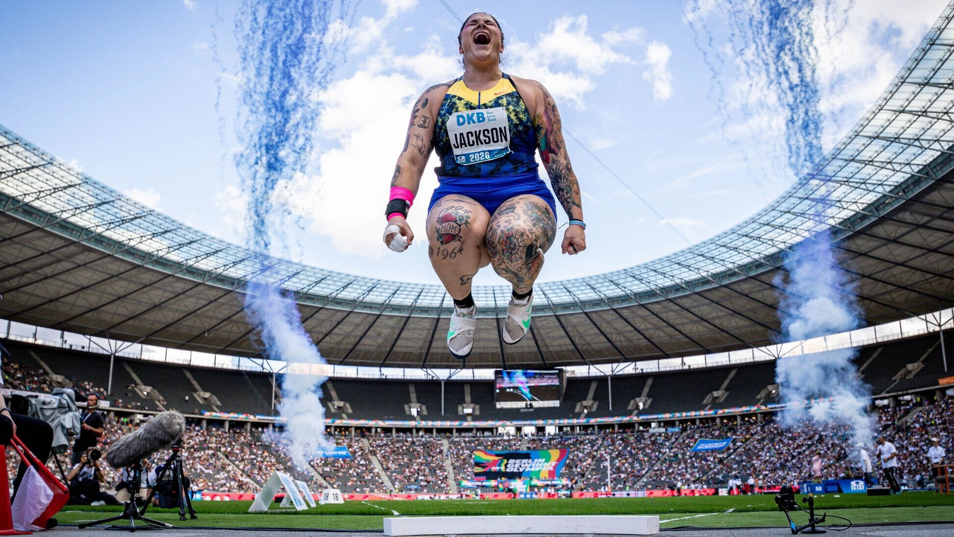 Die US-Kugelstoßerin Chase Jackson springt im einem Stadion in die Luft, während hinter ihr zwei Rauchsäulen aufsteigen.
