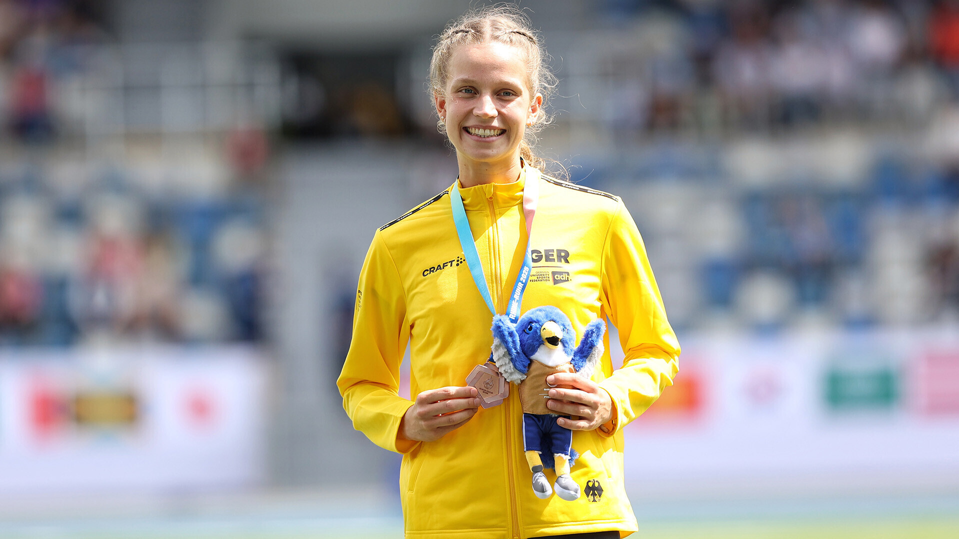 Adia Budde in der gelben Jacke auf dem Podium mit Medaille um den Hals und Maskottchen in der Hand